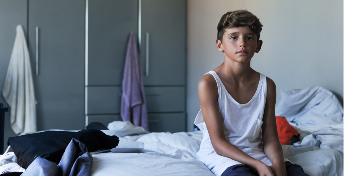 Image of a boy sitting on edge o bed,alone and looking bored.