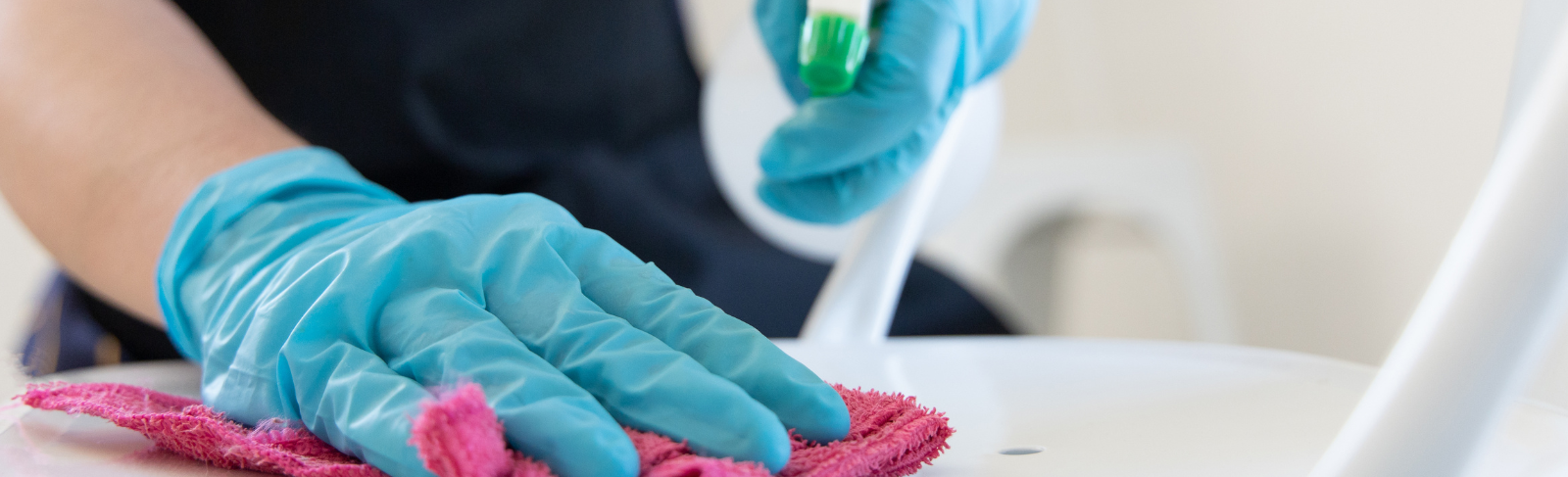 Image of hands wearing latex gloves. One hand is holding a spray bottle, the other is wiping a cloth on a seat.