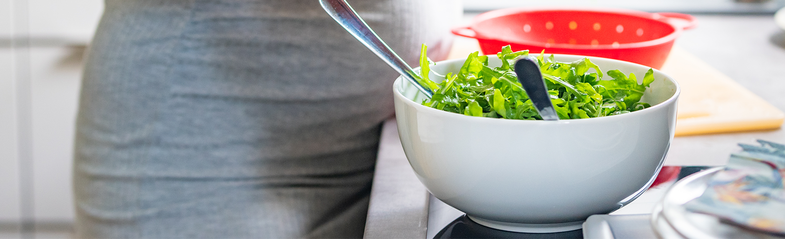 Bowl of salad with a red strainer behind and a person in a grey dress.