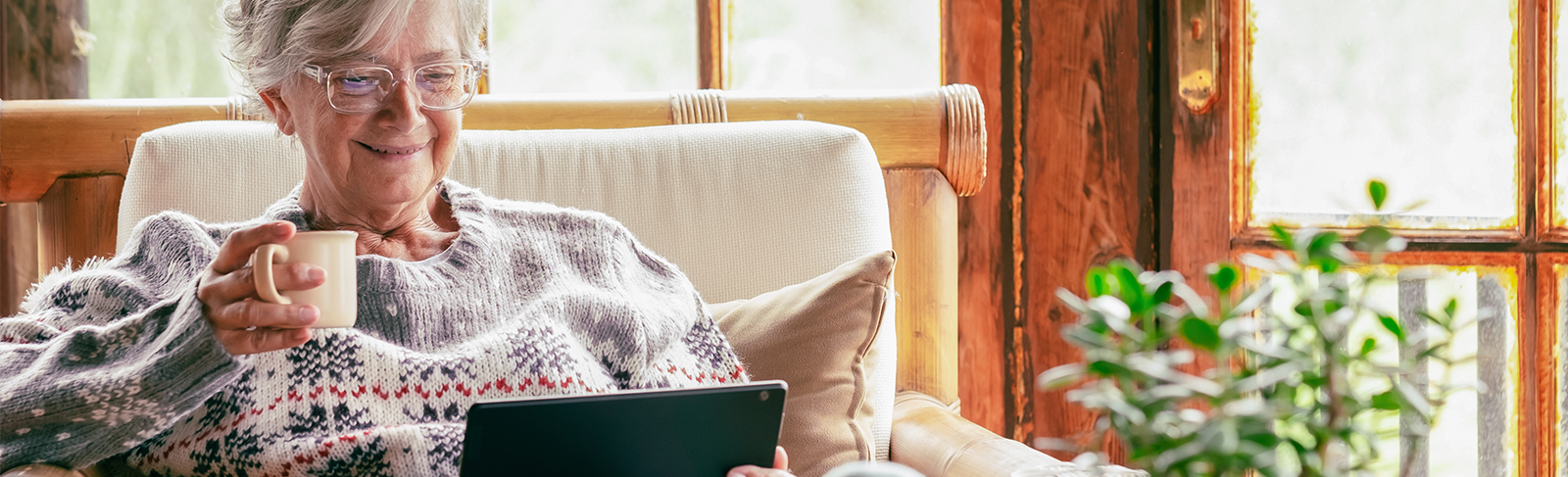 Older adult sitting in a chair with a coffee mug looking at a tablet.