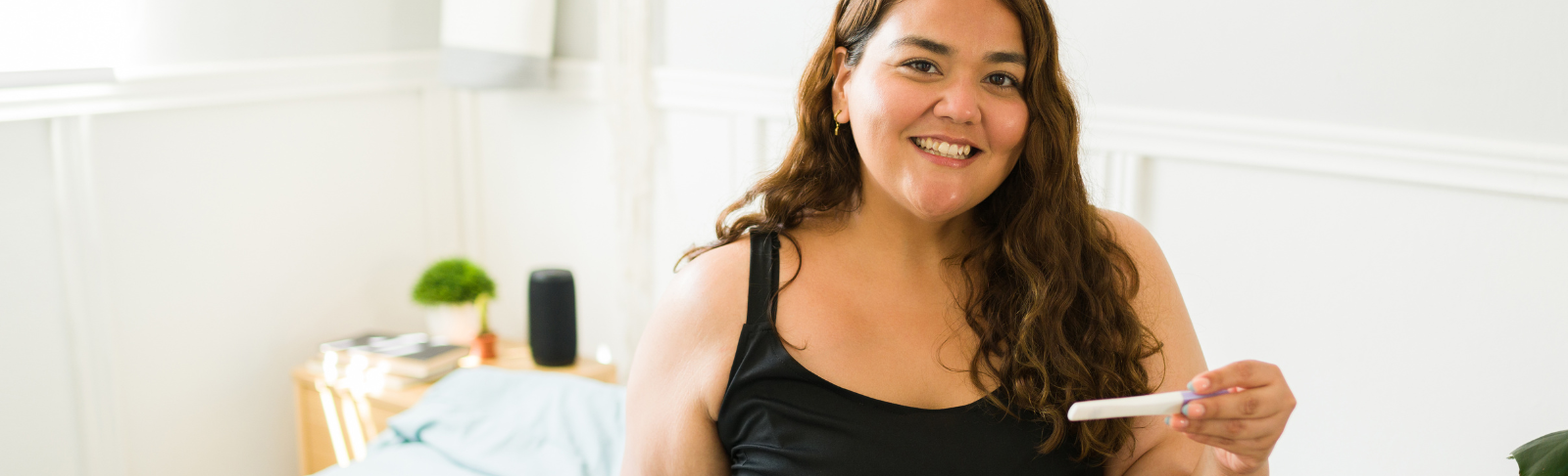 Female-presenting individual sitting on bed, smiling, holding pregnancy test