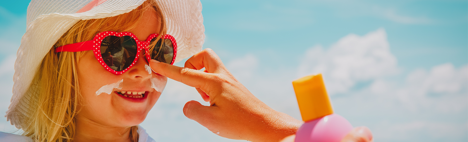 Child wearing a white hat with red glasses having sunscreen put on