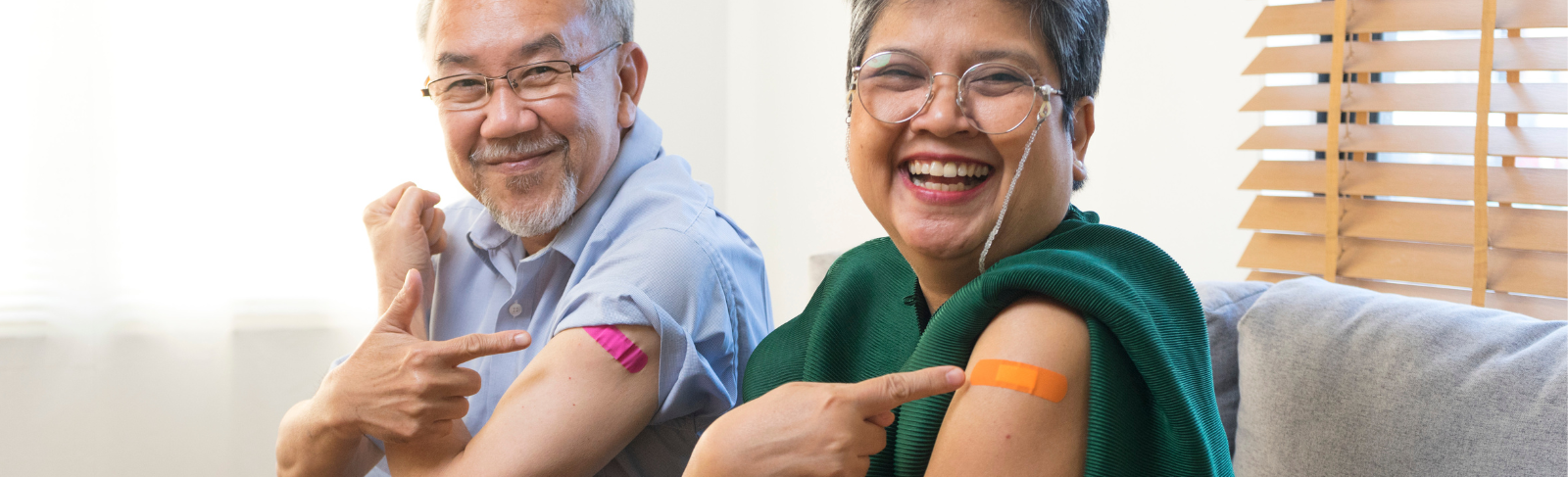 Image of two people sitting on a couch, smiling and pointing to a bandage on their arm