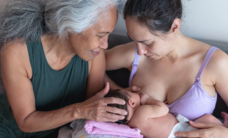 Woman breastfeeding her baby, supported by caring older adult.