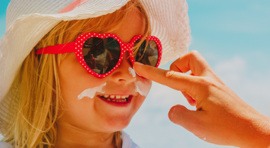 A child wearing wide-brimmed hat and sunglasses, having sunscreen applied to nose.
