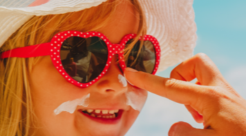 Little girl wearing hat and sunglasses; adult hand applying sunscreen to her nose