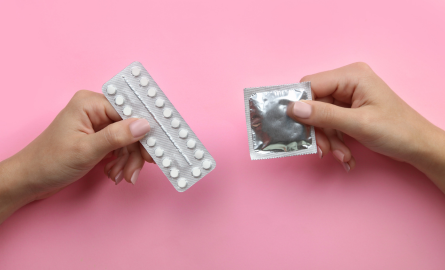 Two hands in front of a pink background, left hand is holding a package of birth control pills, right hand is holsing a condom in its packaging