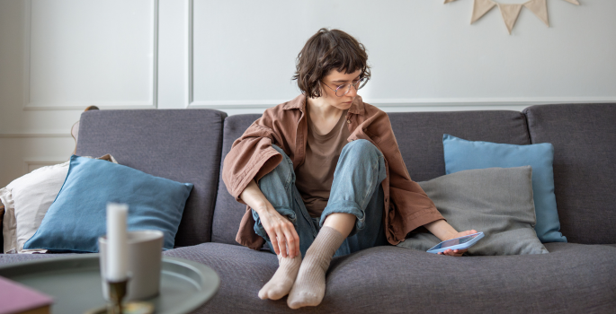 Image of young adult alone on couch, reaching to grab cell phone