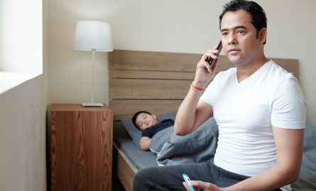 Male-presenting individual sitting at edge of bed and holding phone to ear, looking distressed. In background, child lays on bed.