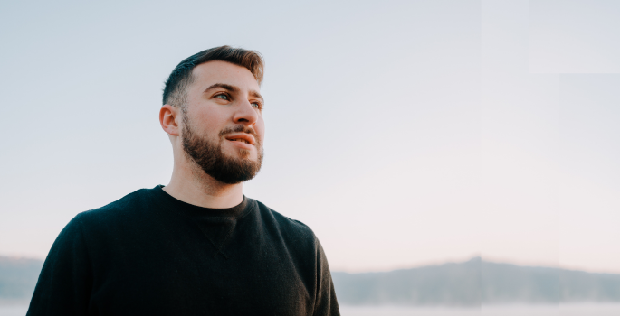 Person outdoors, looking into the distance. A tree-lined lake is far in the background.