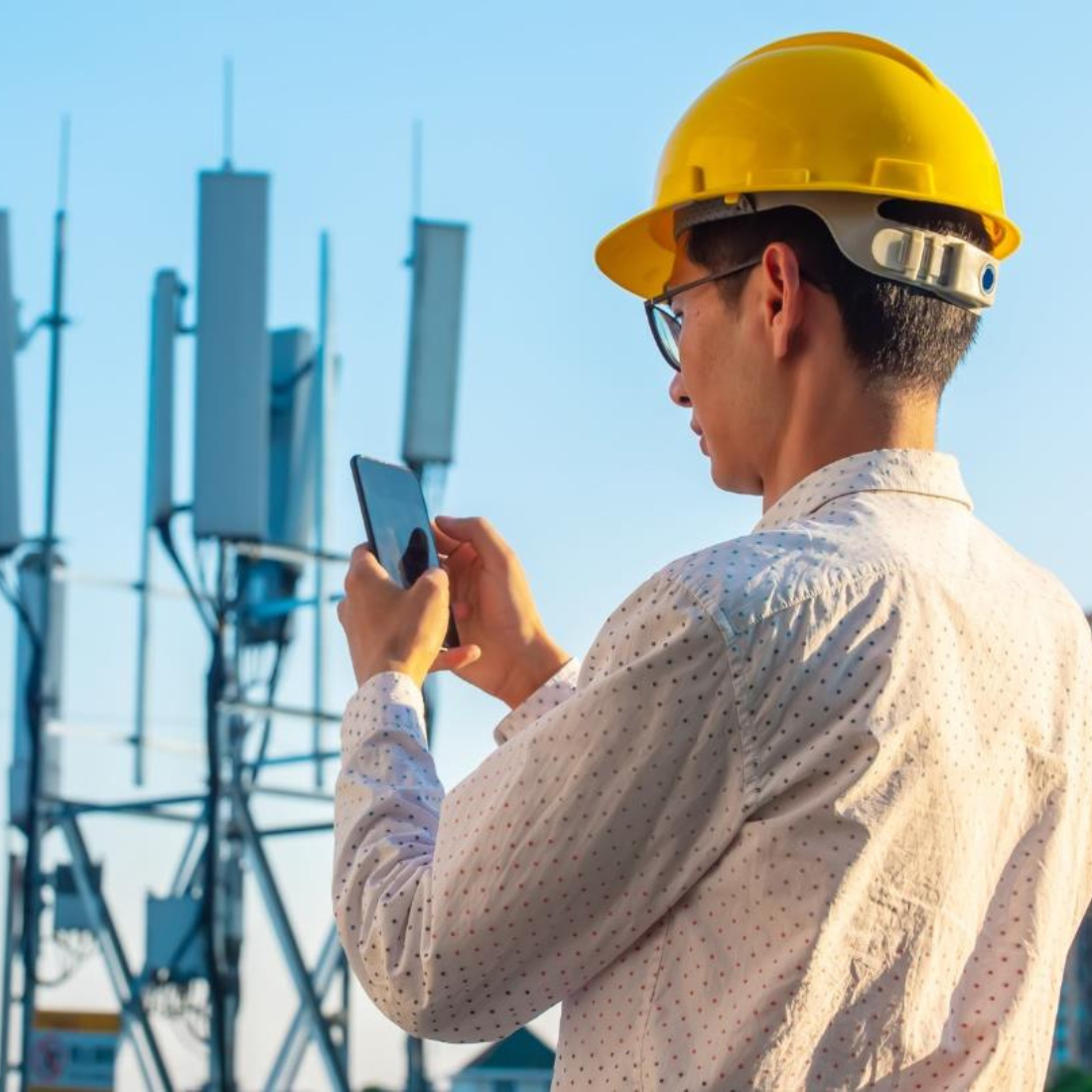 Image of a person wearing a hard hat, holding up a cell phone near industrial towers.