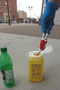 Image of tongs being used to place a sharp in a biohazard container. Hands using the tongs are wearing gloves. Next to the container is a plastic pop battle with cap.