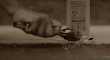 Image of a hand using tongs to pick up a glass pipe from the ground. In the background, a biohazard container.