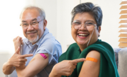Two adults smiling and pointing to bandage on upper arms