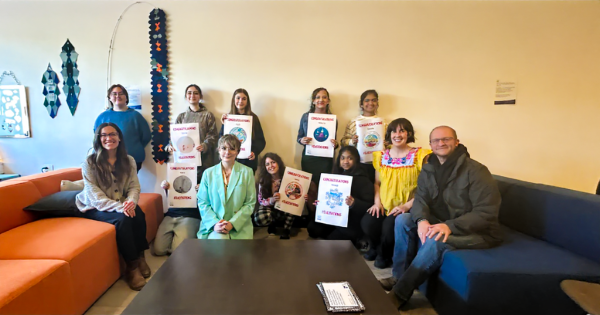 Group photo from Sticker Design Reveal Party Group of youth standing or crouching in front of a wall, each holding a unique picture. Smiling adults sit next to them on either side. On ie closer to the middle.