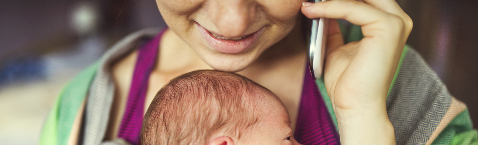 Close up image of someone smiling while holding phone to ear and carrying baby in baby carrier 