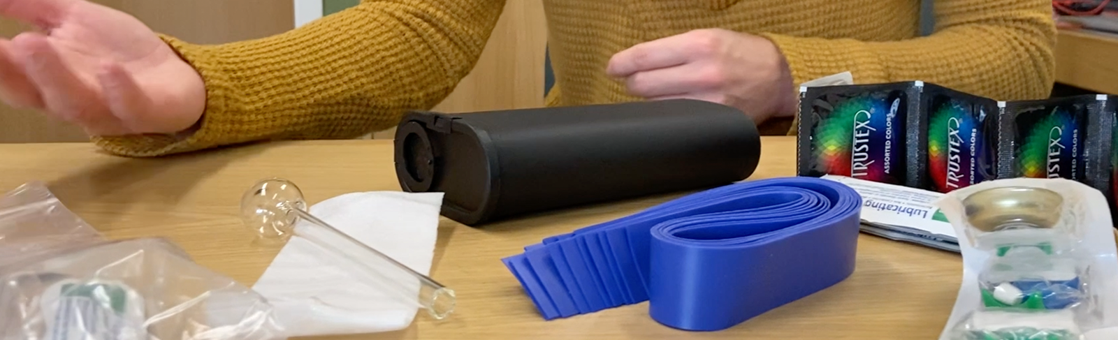Harm reduction items like condoms and pipes on a table with a person's arm's behind. 