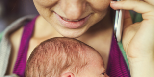 Close-up image of a smiling parent holding phone to ear and baby resting against chest while in carrier