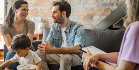 Young couple seated in living room, having conversation with another adult while toddler plays.