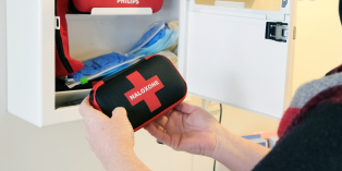 Hands holding a naloxone kit outside an open, wall-mounted First Aid cupboard