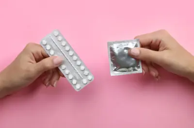 Two hands in front of a pink background, left hand is holding a package of birth control pills, right hand is holsing a condom in its packaging 