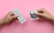 Two hands in front of a pink background, left hand is holding a package of birth control pills, right hand is holsing a condom in its packaging 