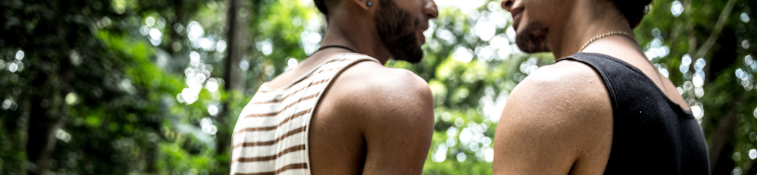 Two males standing intimately, facing one another against forested background